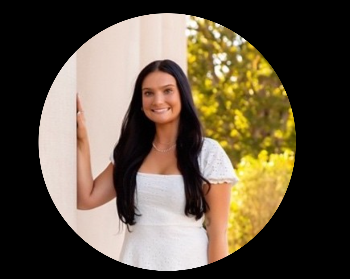Woman in white dress leaning against a white pillar, smiling outside with green trees.