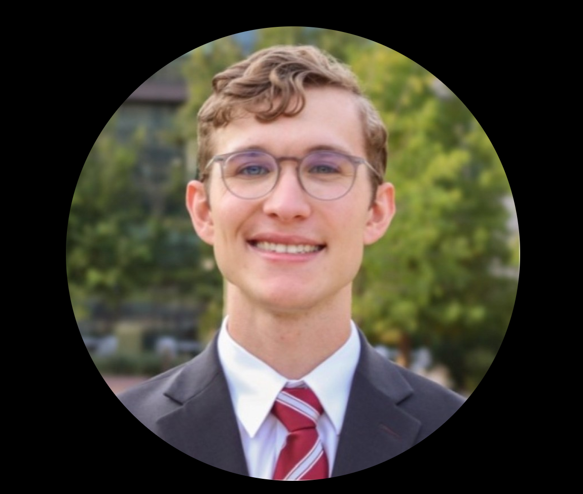 Man with glasses and tie smiling, wearing a suit, outdoors.
