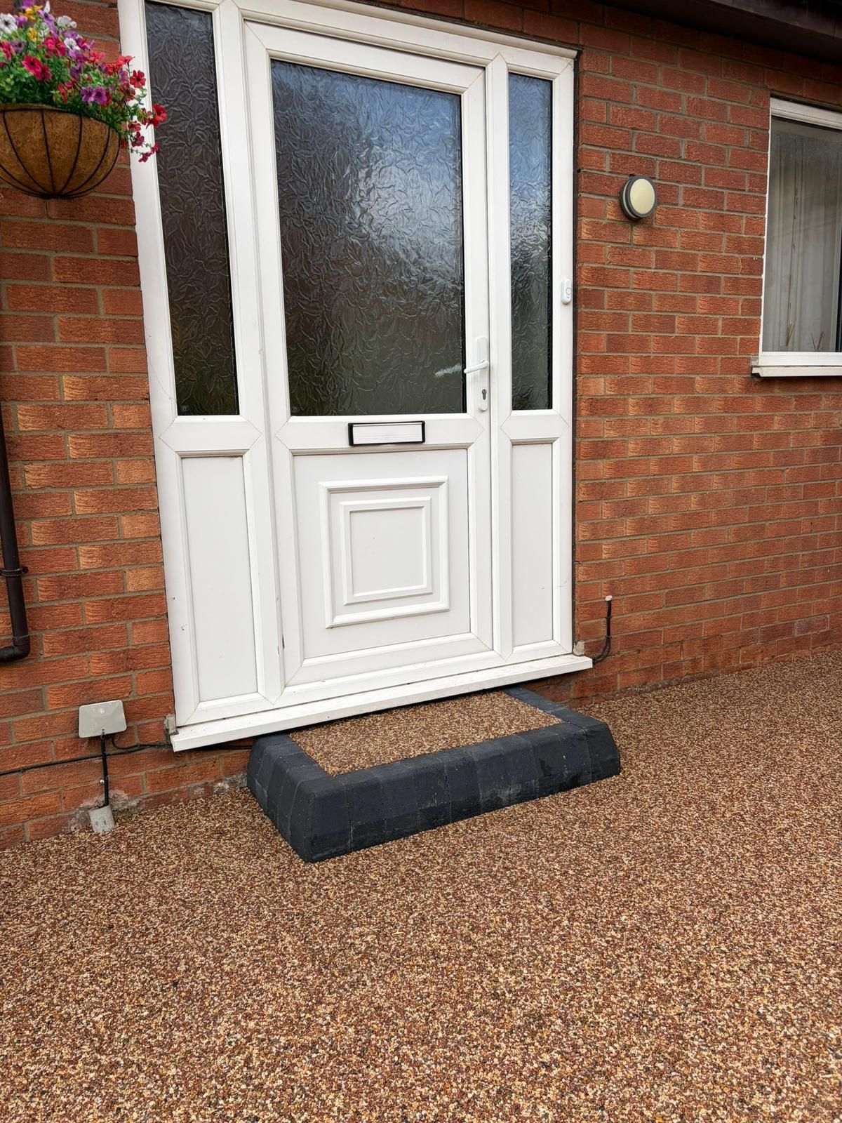 A white front door with side panels, a dark step, and a hanging flower basket on a red brick house with a gravel ground.