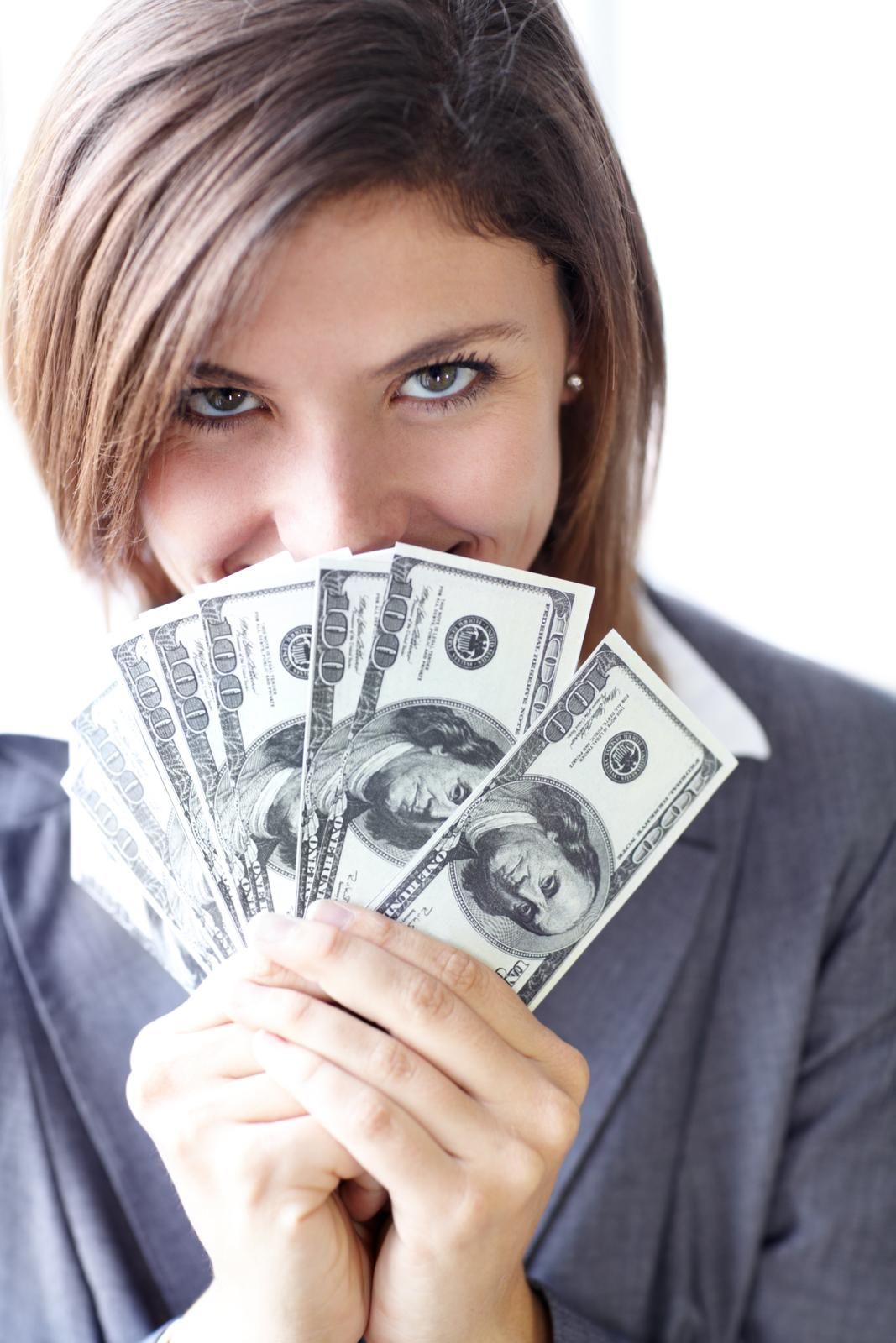 Junge Frau versteckt grinsend ihr Gesicht hinter einigen 100 US-Dollar Noten
