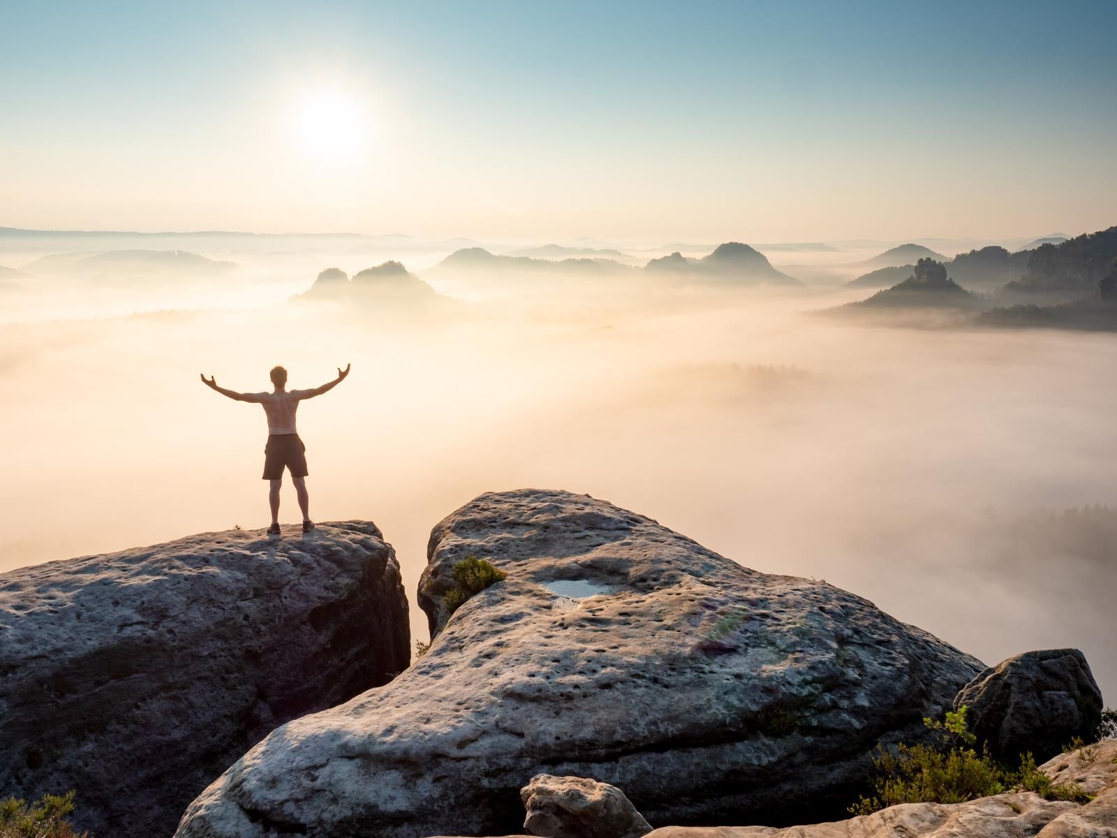 Symbolbild für Freiheit, Mann steht auf den Bergen oberhalb der Wolken und fühlt sich frei mit Blick auf die Sonne.