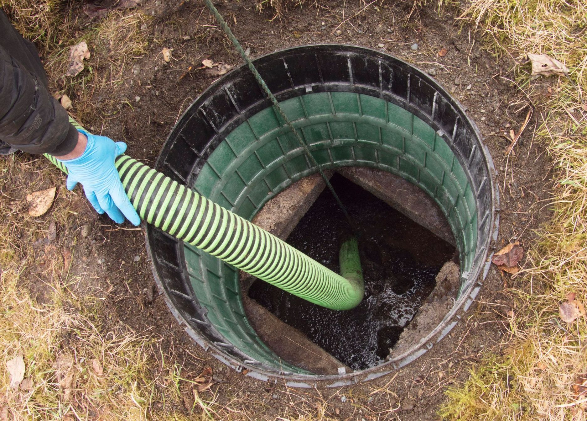 Person with blue gloves guiding a striped green hose into an open septic tank.