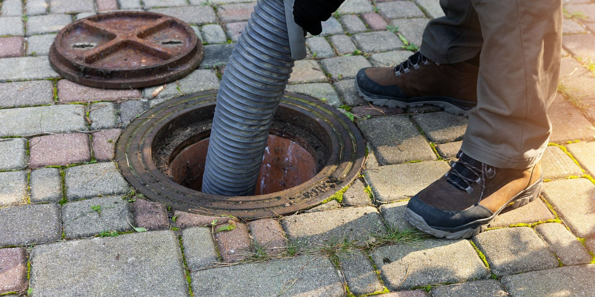 A person is pumping sewage from a manhole. A large hose descends into the manhole while the cover rests nearby on a brick-paved ground.