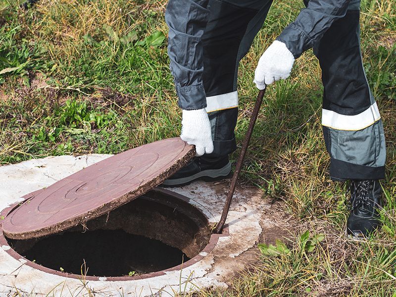 Person in work clothes lifting a manhole cover with a hook in a grassy area.