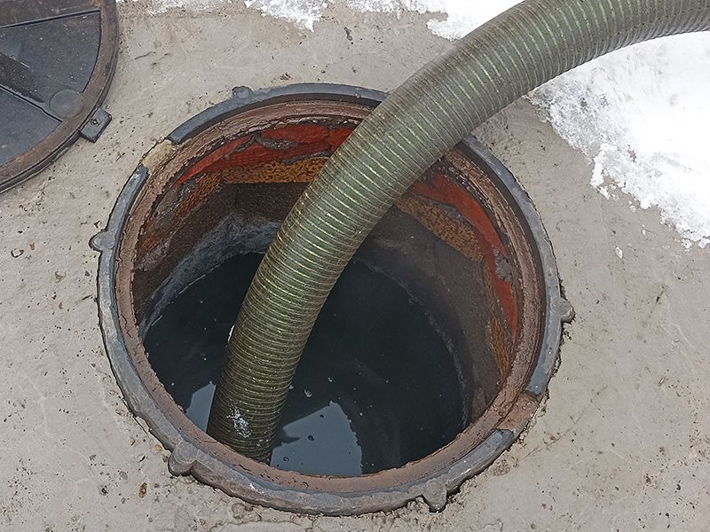 A septic tank being pumped out by a hose. The open tank is round with dark liquid inside, and the hose is green.