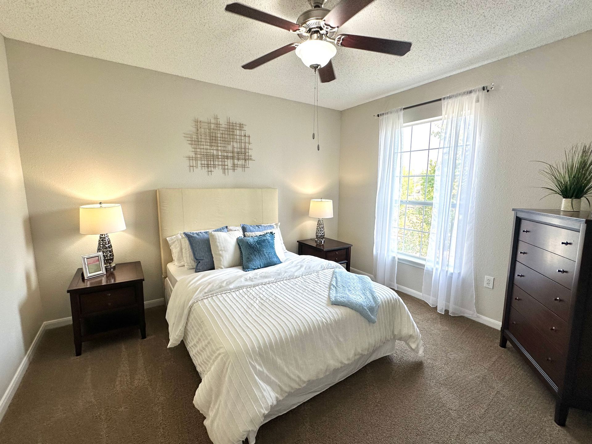 Bedroom with bed, nightstands, dresser, window, and ceiling fan. Beige walls, brown carpet, and white bedding.