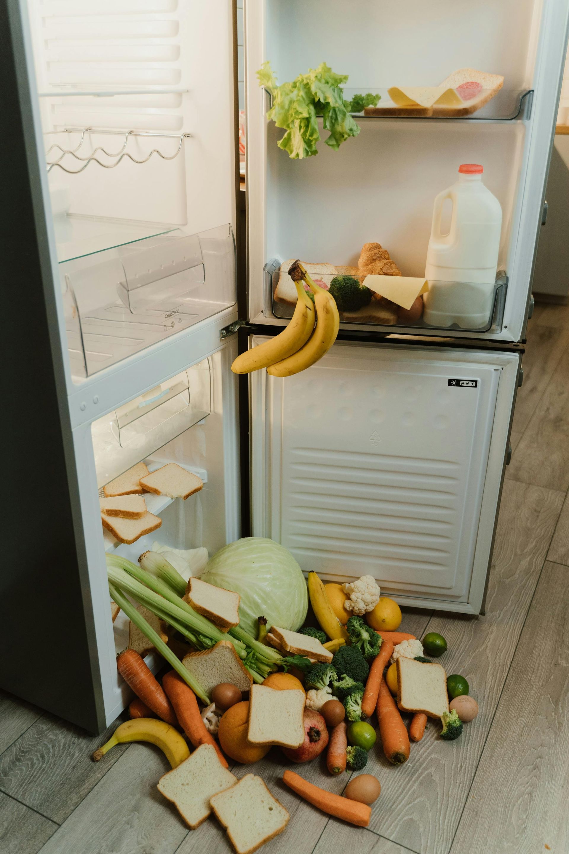 Open fridge with spilled fruits and vegetables on the floor; bananas hang from the shelf.