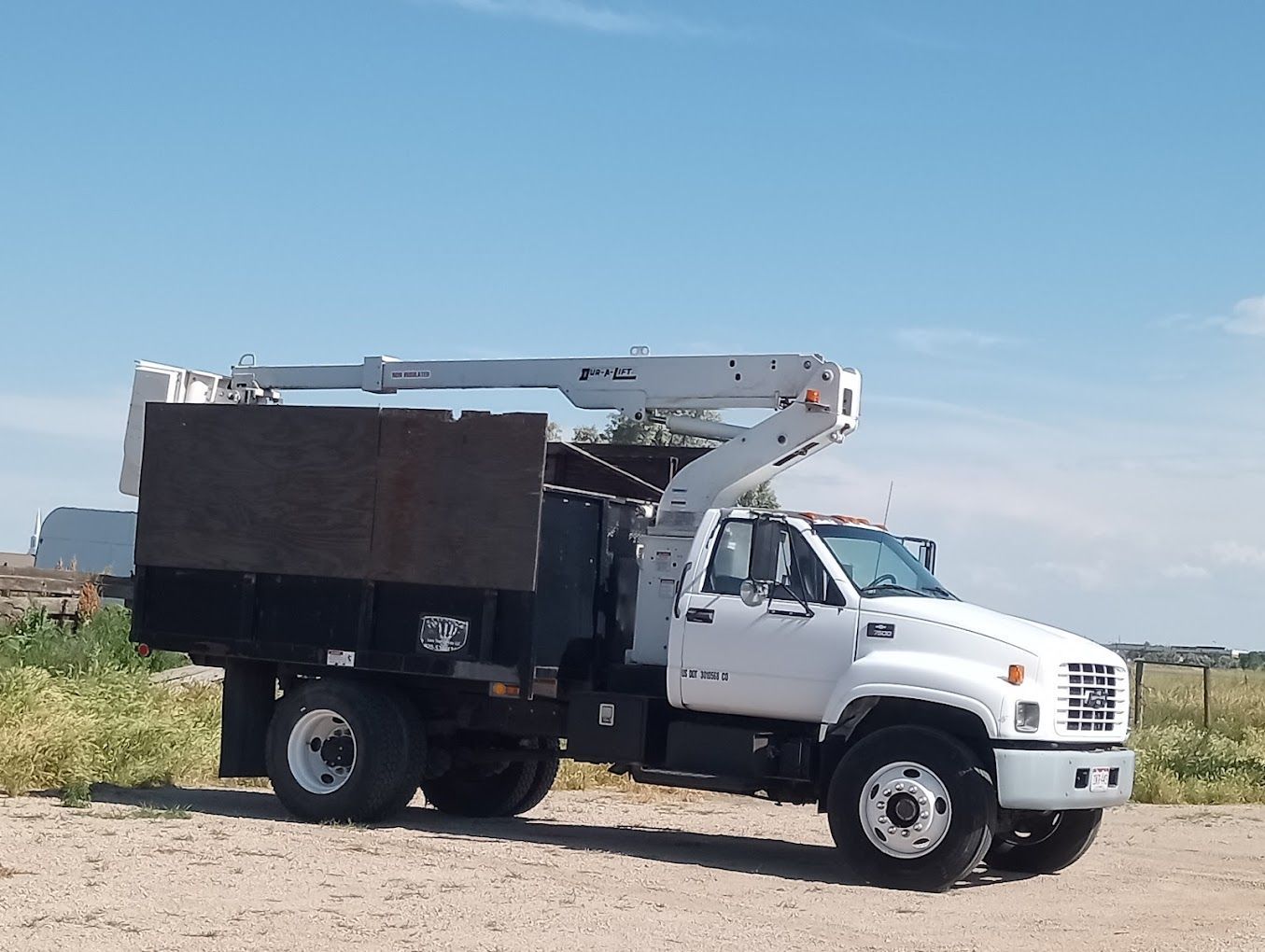 White tree trimming truck with an extended boom arm on a dirt road under a blue sky.