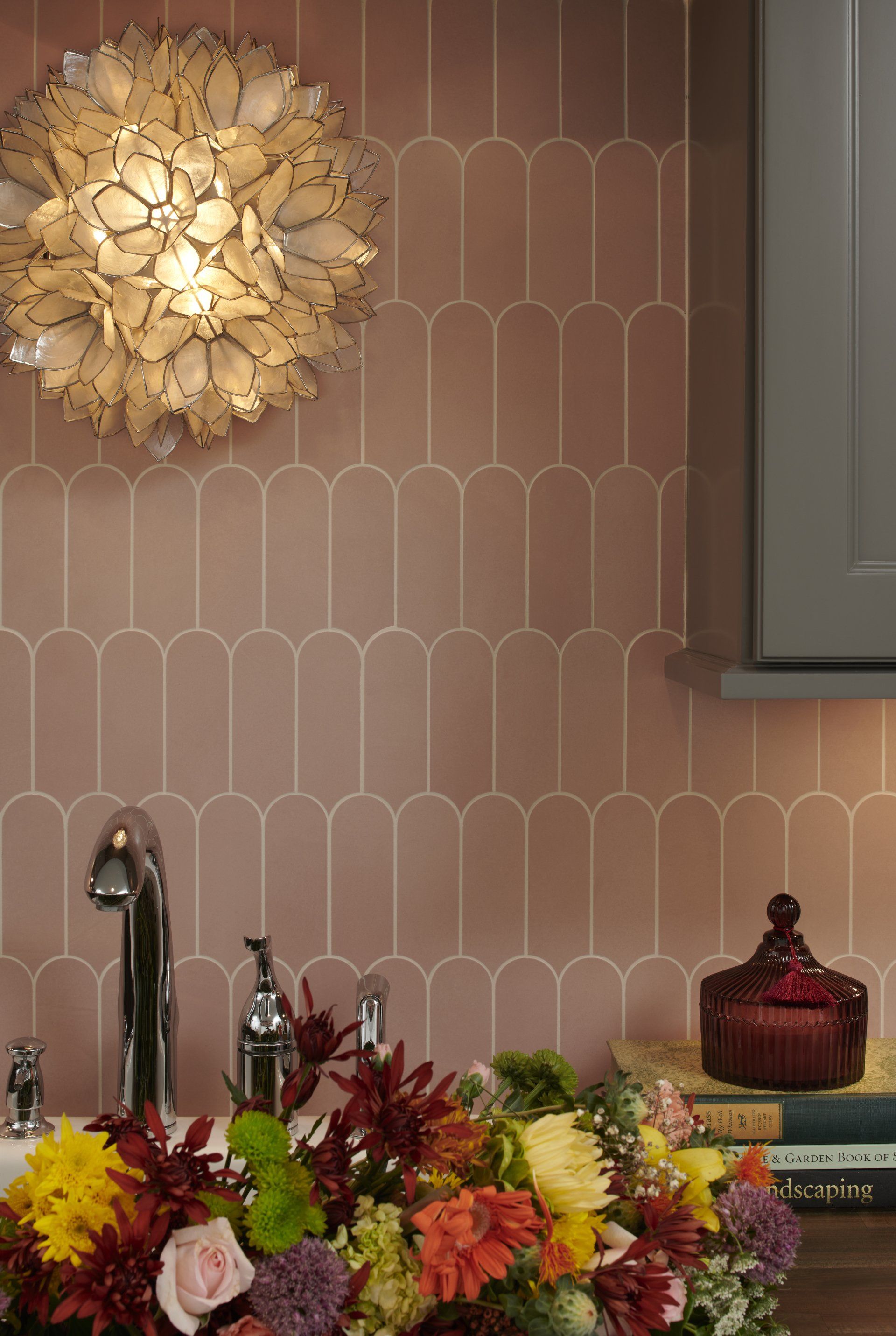 A kitchen with pink tiles , a sink , a chandelier and flowers on the counter.