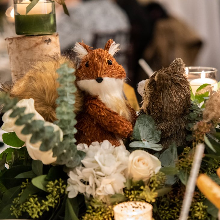 A stuffed fox is sitting on a table surrounded by flowers and candles