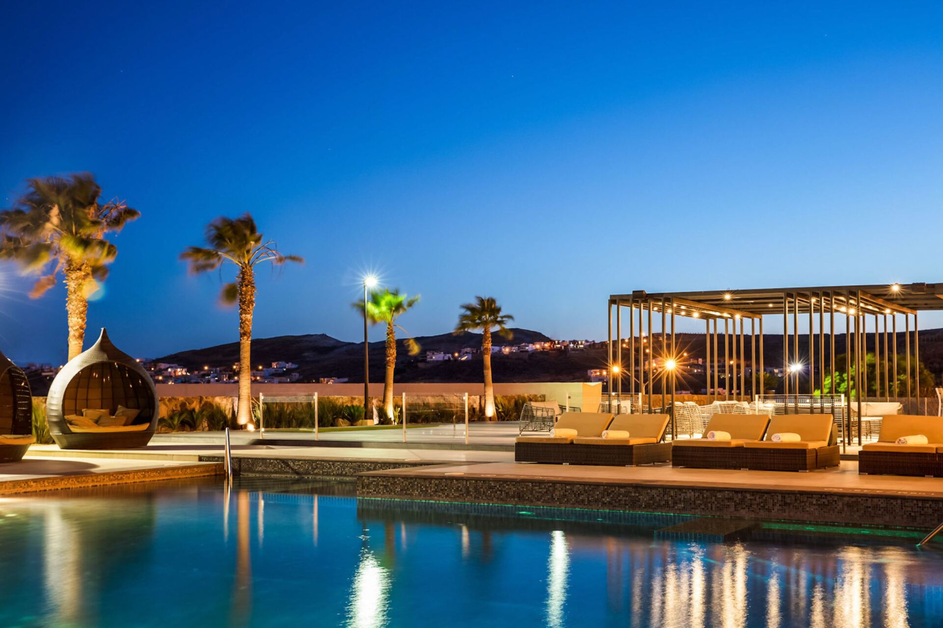 A large swimming pool is lit up at night with palm trees in the background.