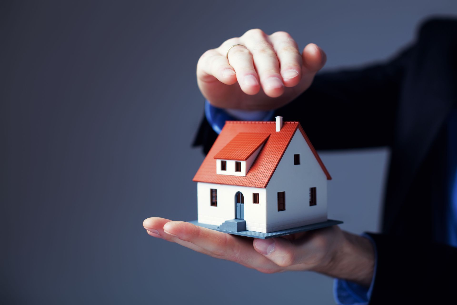 Person in a suit holding a miniature house, with one hand above to protect it.