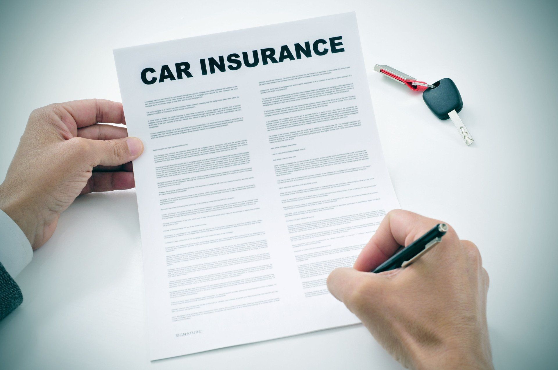 Person signing a car insurance document; car keys rest nearby on a white surface.