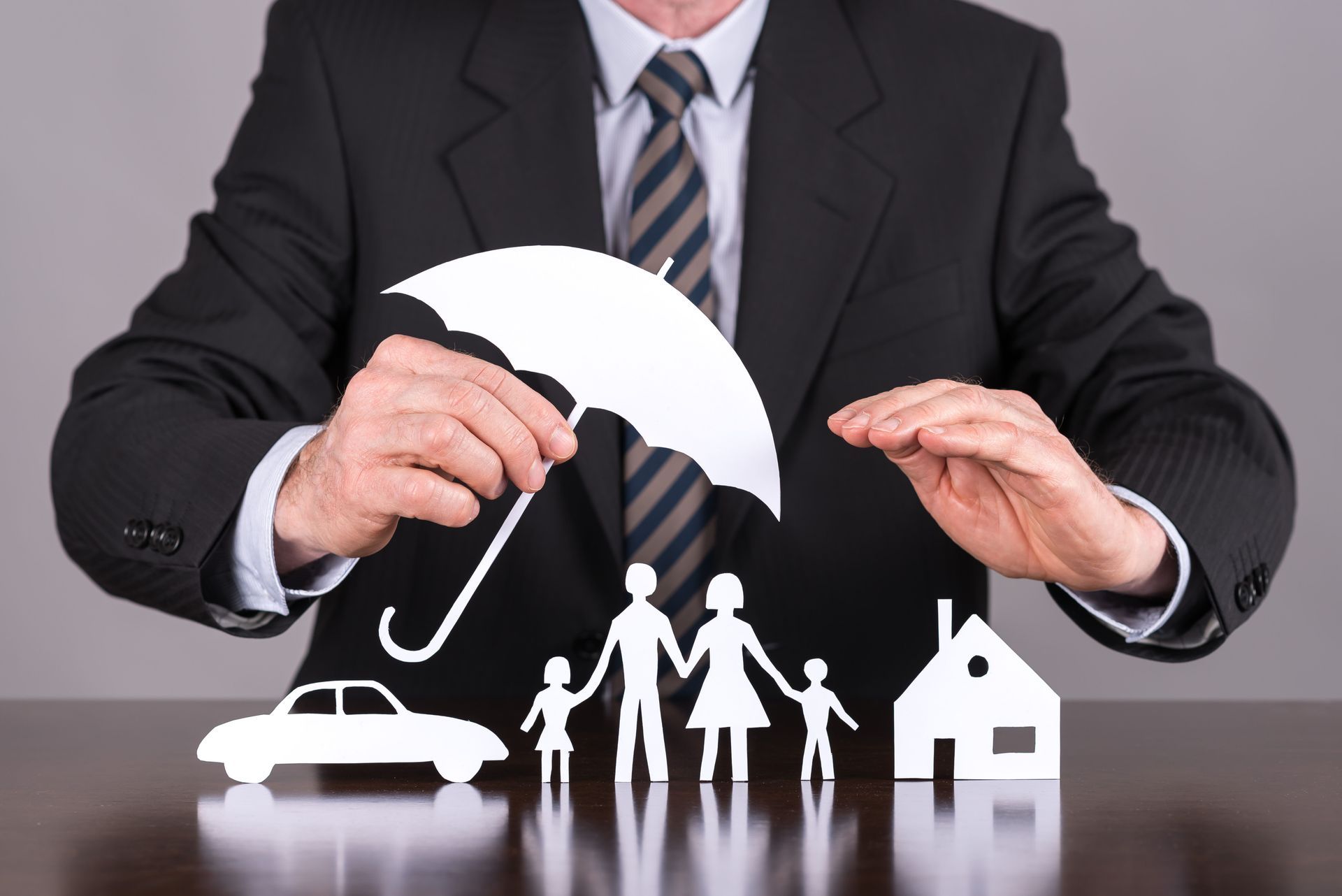 Man in a suit holding a paper umbrella above paper cutouts of a car, family, and house, representing insurance coverage.