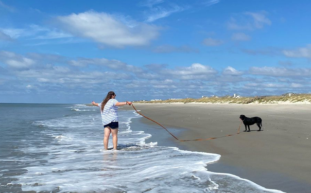 Person with outstretched arms holds a stick, dog stands on beach. Ocean and blue sky with clouds in background.