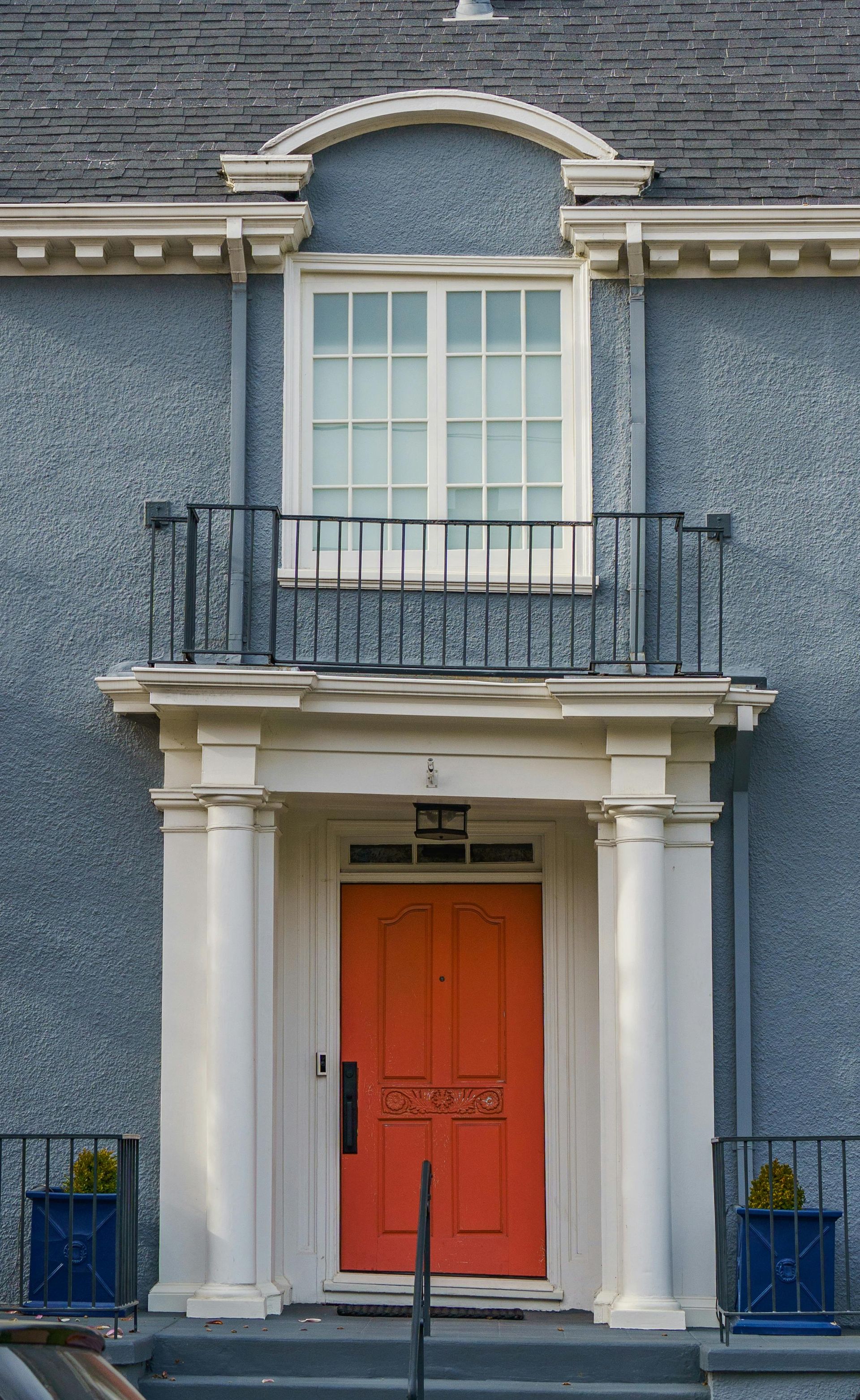A vibrant orange double front door set within a grey stone building, framed by white columns and a decorative balcony.