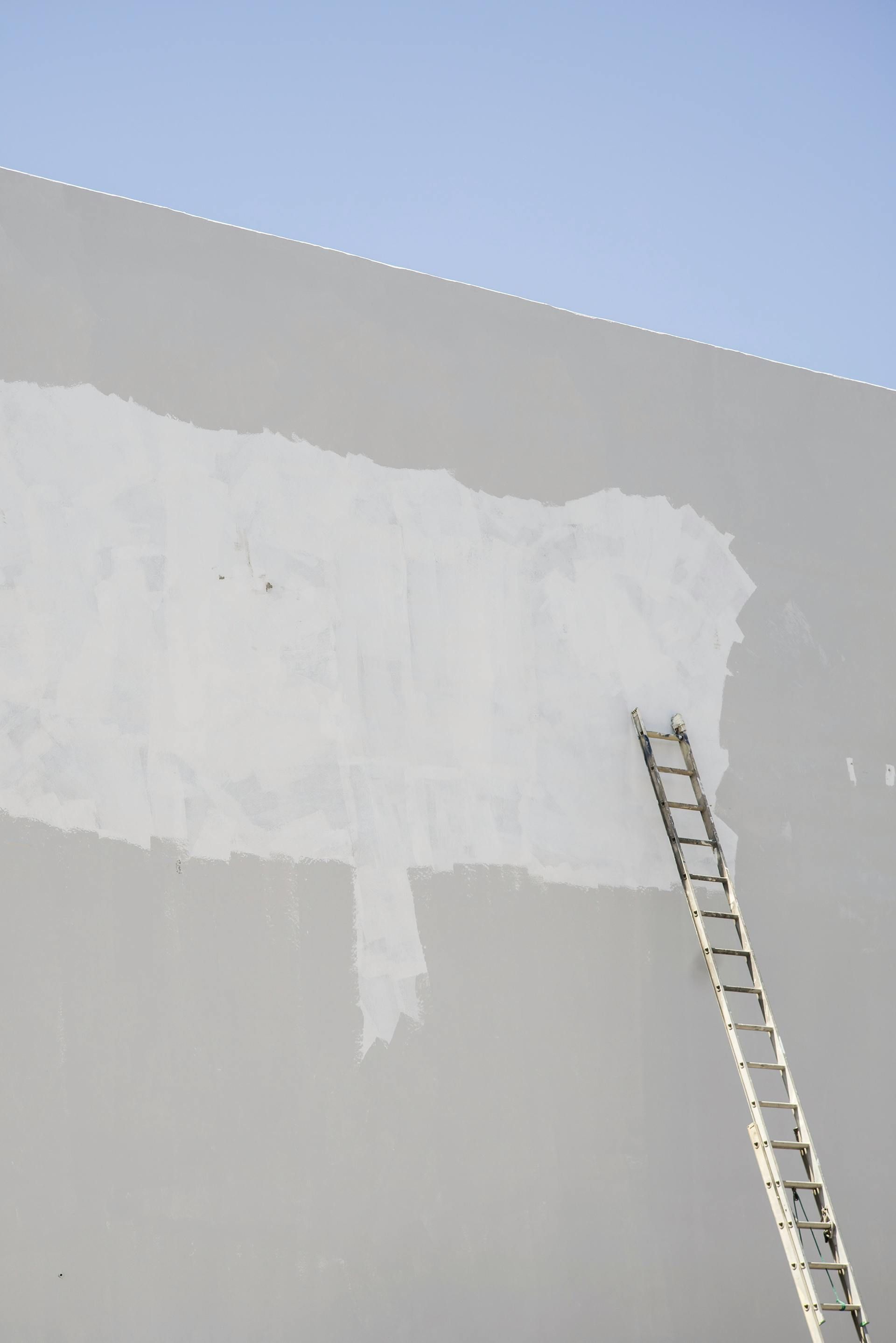 An extension ladder leans against a high grey wall with a large, roughly painted white patch in the center.