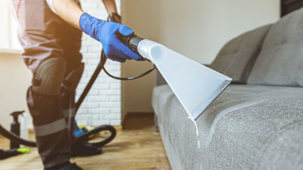 A man is cleaning a couch with a vacuum cleaner.