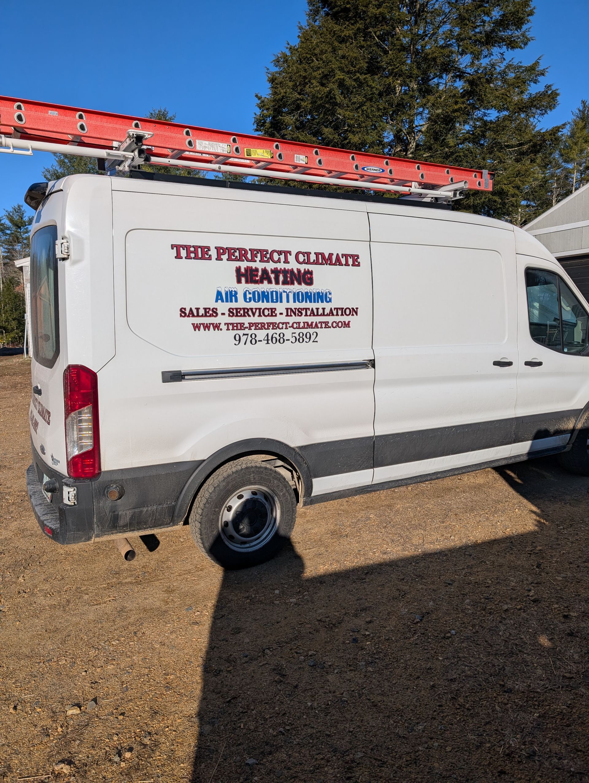 A white van with a ladder on top of it is parked in a gravel lot.