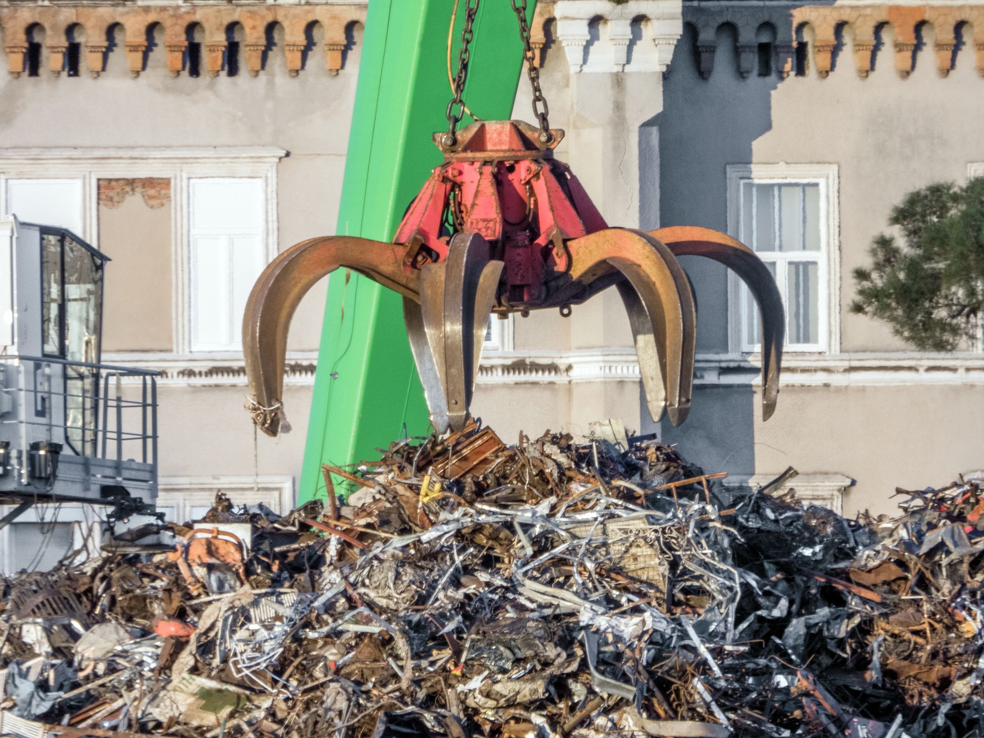 Industrial claw grabbing metal pile for scrap metal recycling near residential building.