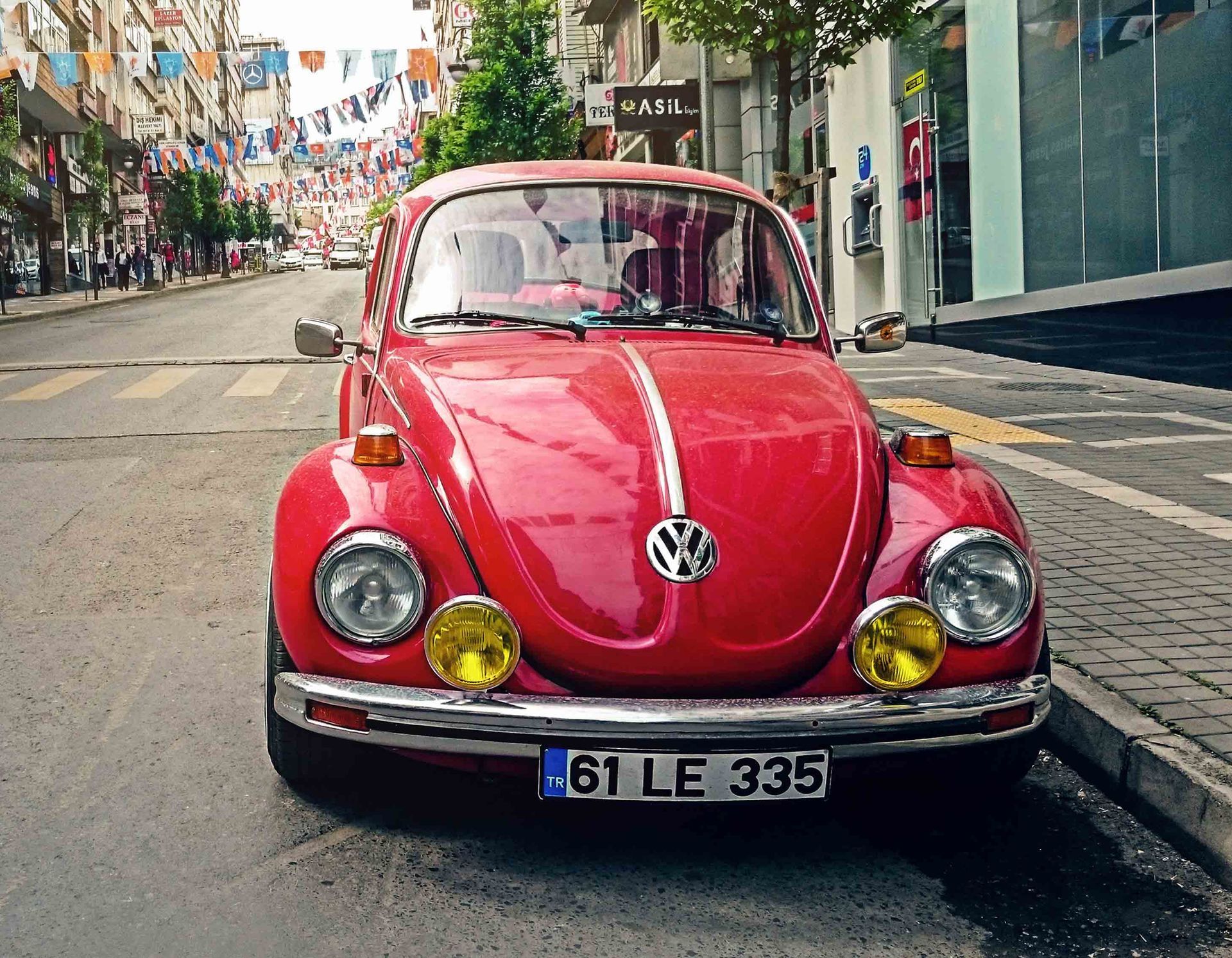Red vintage Volkswagen Beetle parked on a street, with colorful flags overhead.