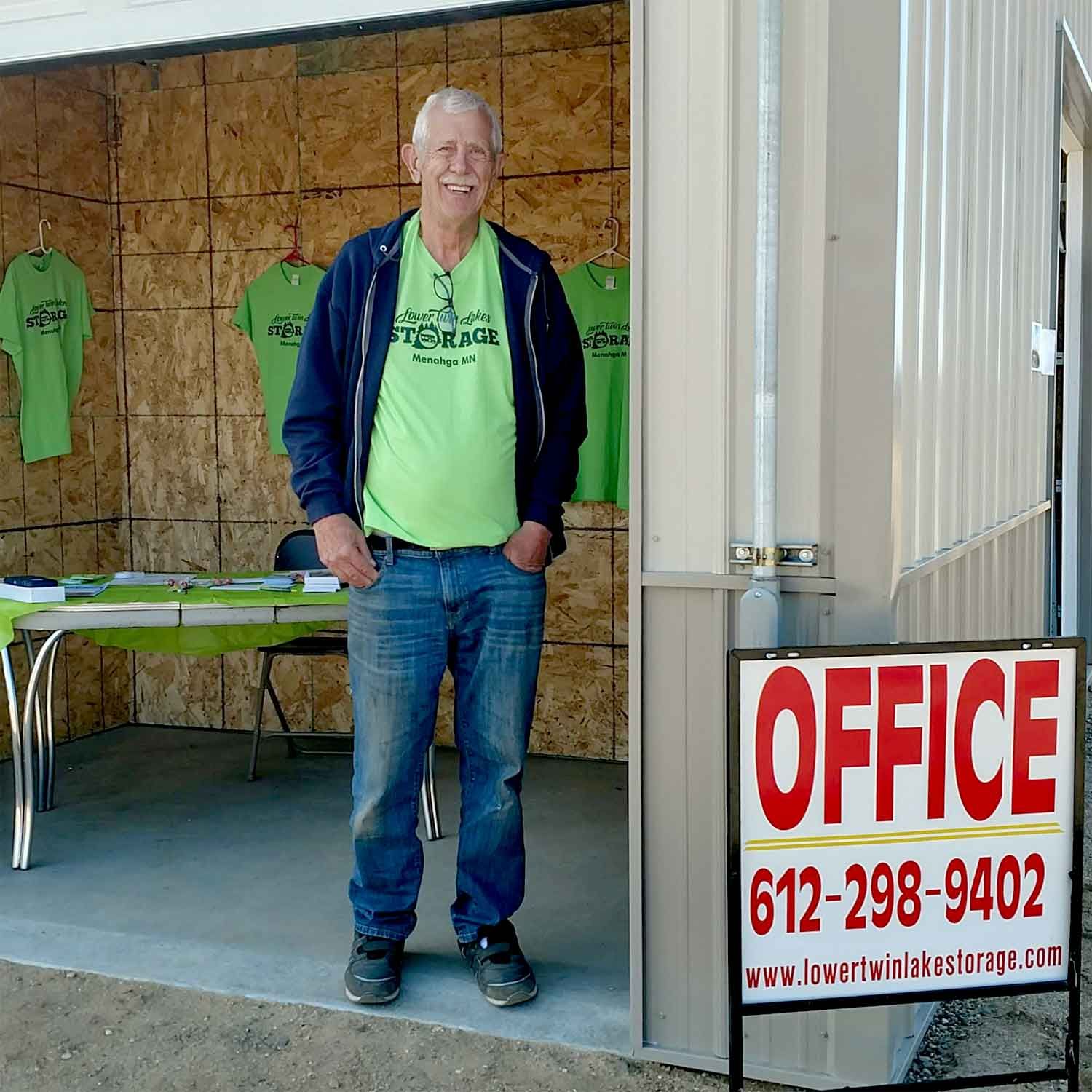 Man standing at office entrance; wearing green shirt, jeans; office sign with phone number.
