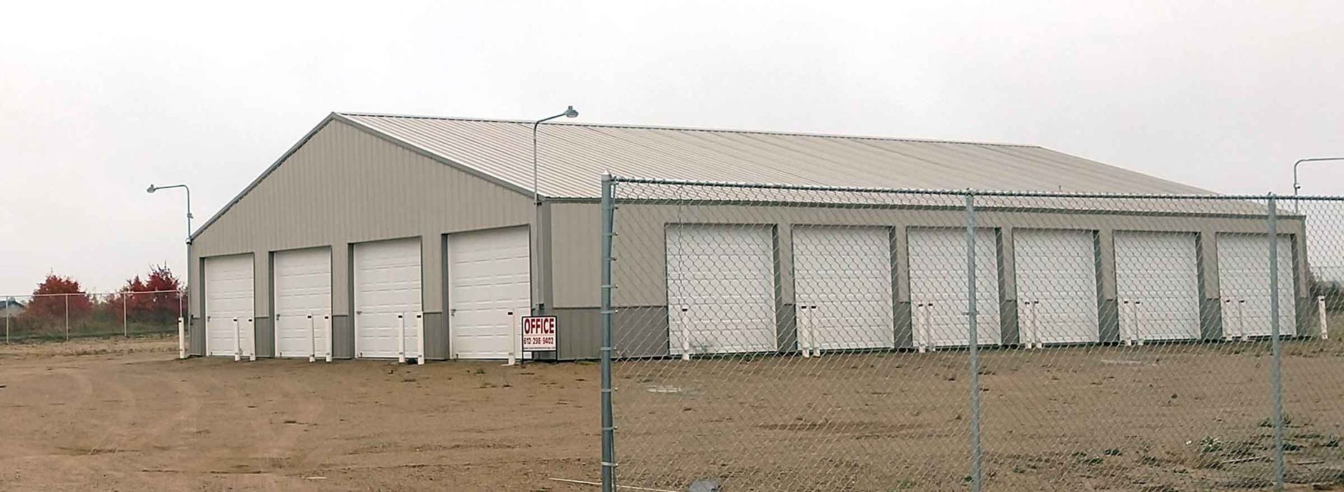 Storage units behind a chain-link fence. Gray building with white garage doors. Brown dirt ground. Overcast sky.