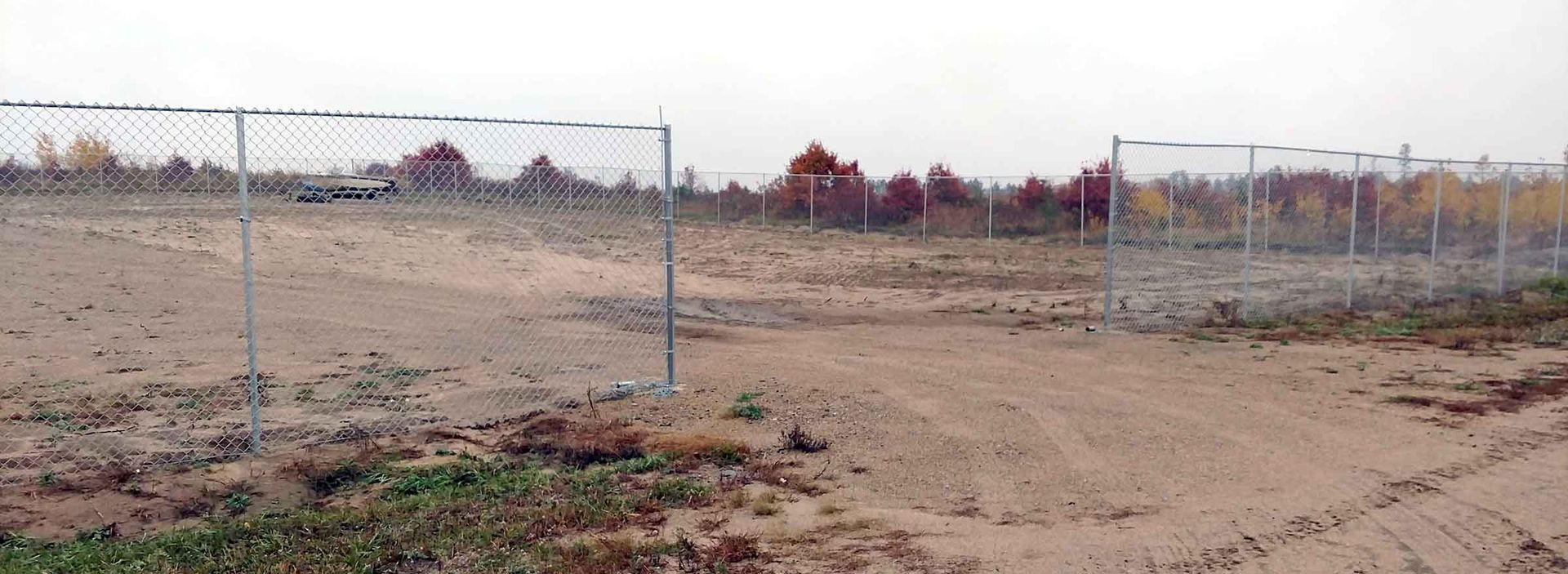 Chain-link fence surrounds a dirt lot. Trees are visible in the distance under a cloudy sky.