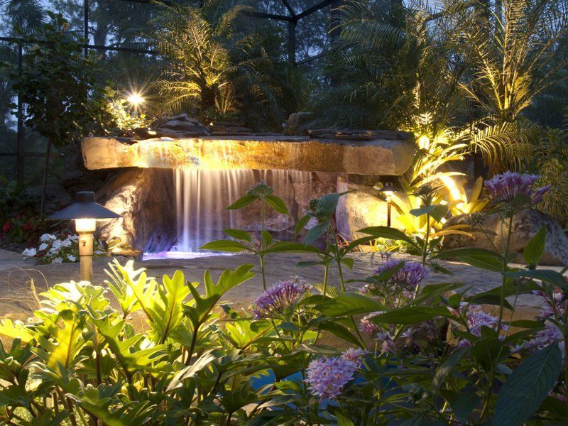 Lit waterfall feature in a garden, illuminated by yellow lights at night, with purple flowers in foreground.