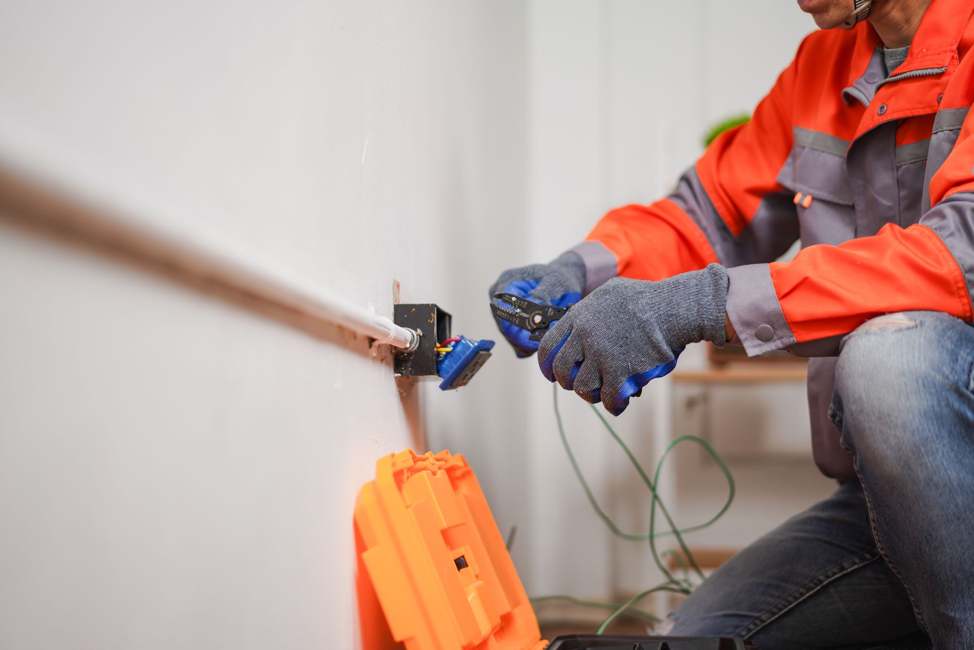 Electrician in orange safety jacket, wiring a wall outlet.