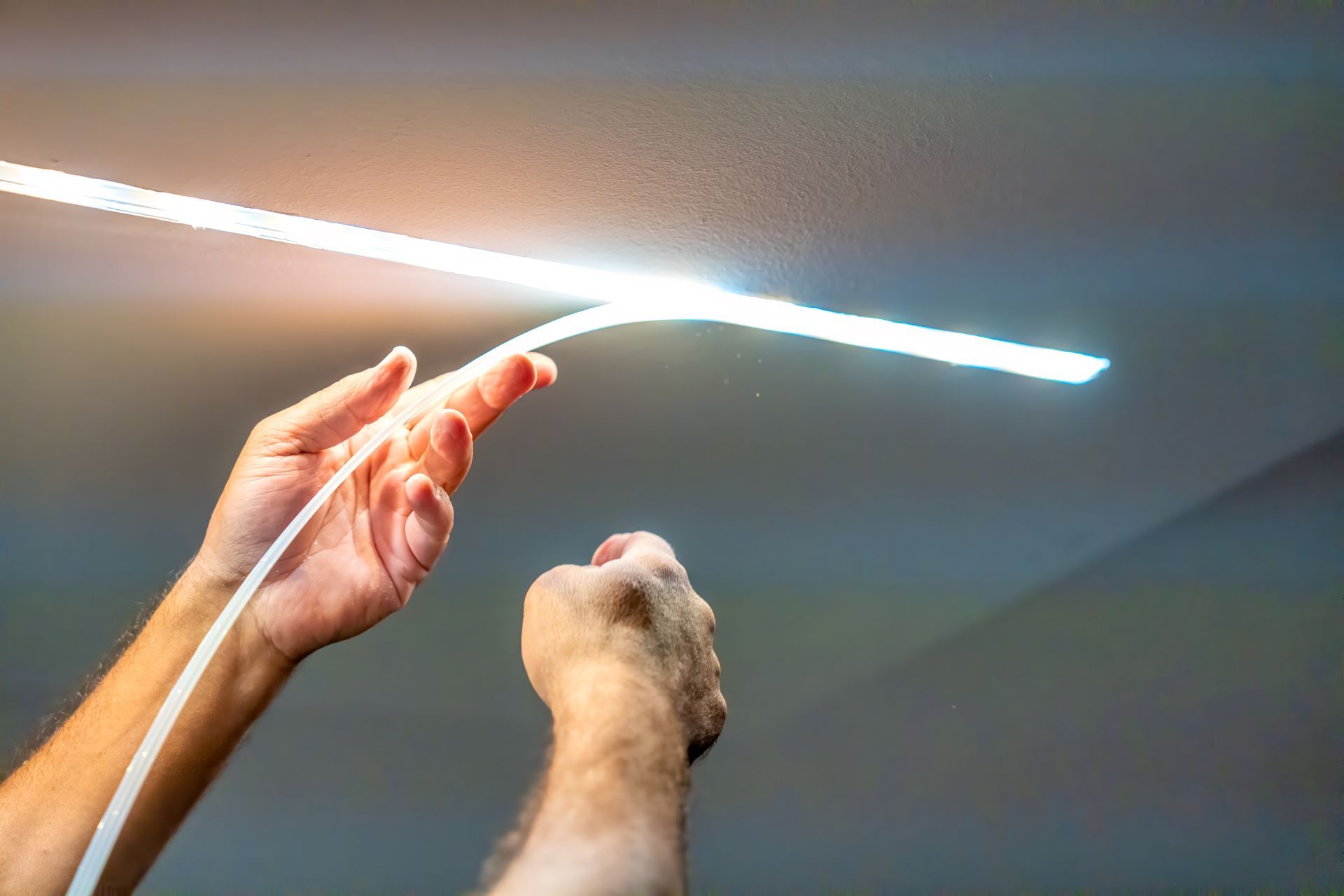 Hands holding a lit, flexible LED light strip against a white ceiling.