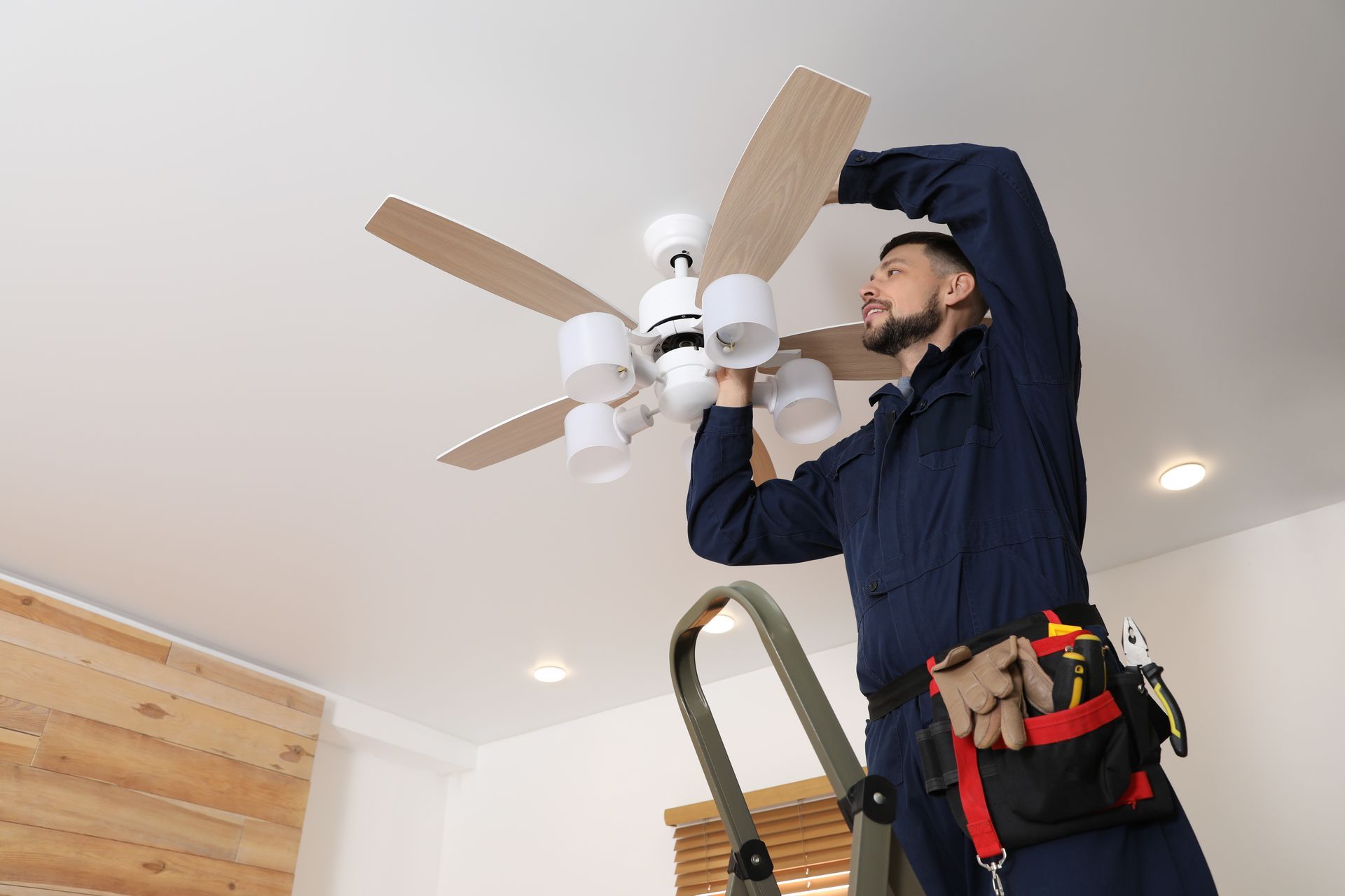Electrician on a ladder installing a ceiling fan with lights in a room with wood paneling.