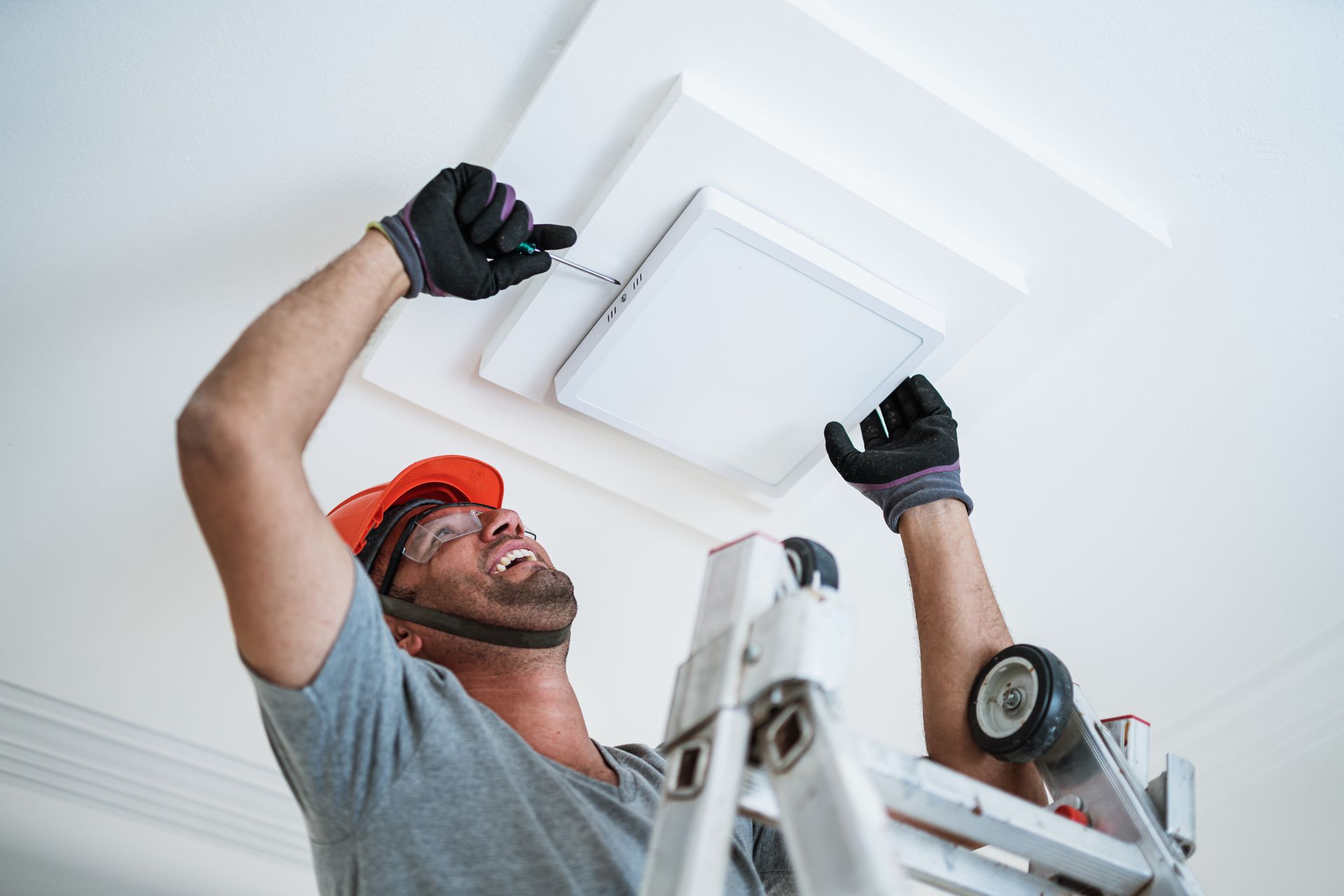 Man in hard hat and gloves on a ladder installing a ceiling light fixture.