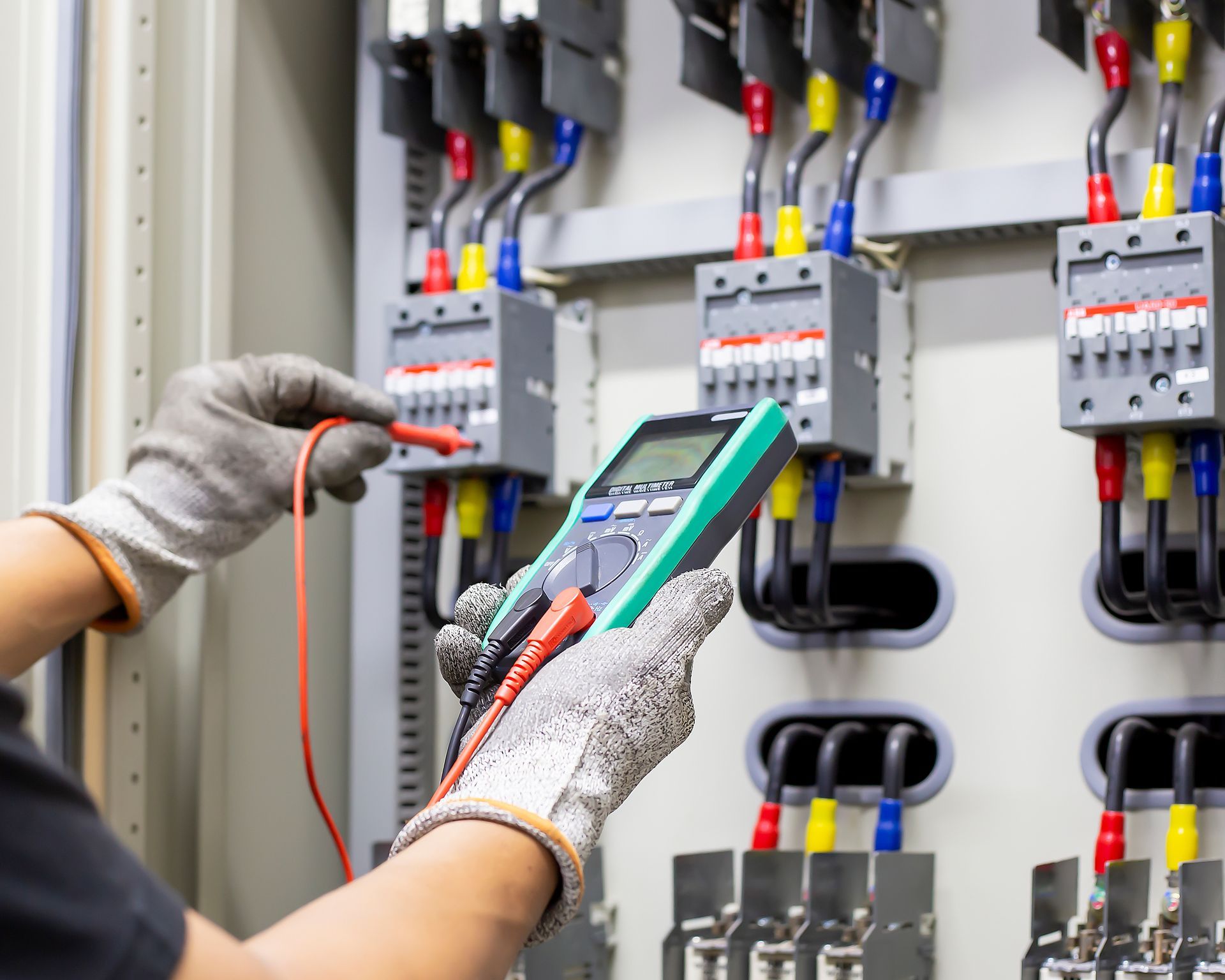 An electrician uses a multimeter to inspect electrical wiring in a panel. Wires are red, blue, and yellow.