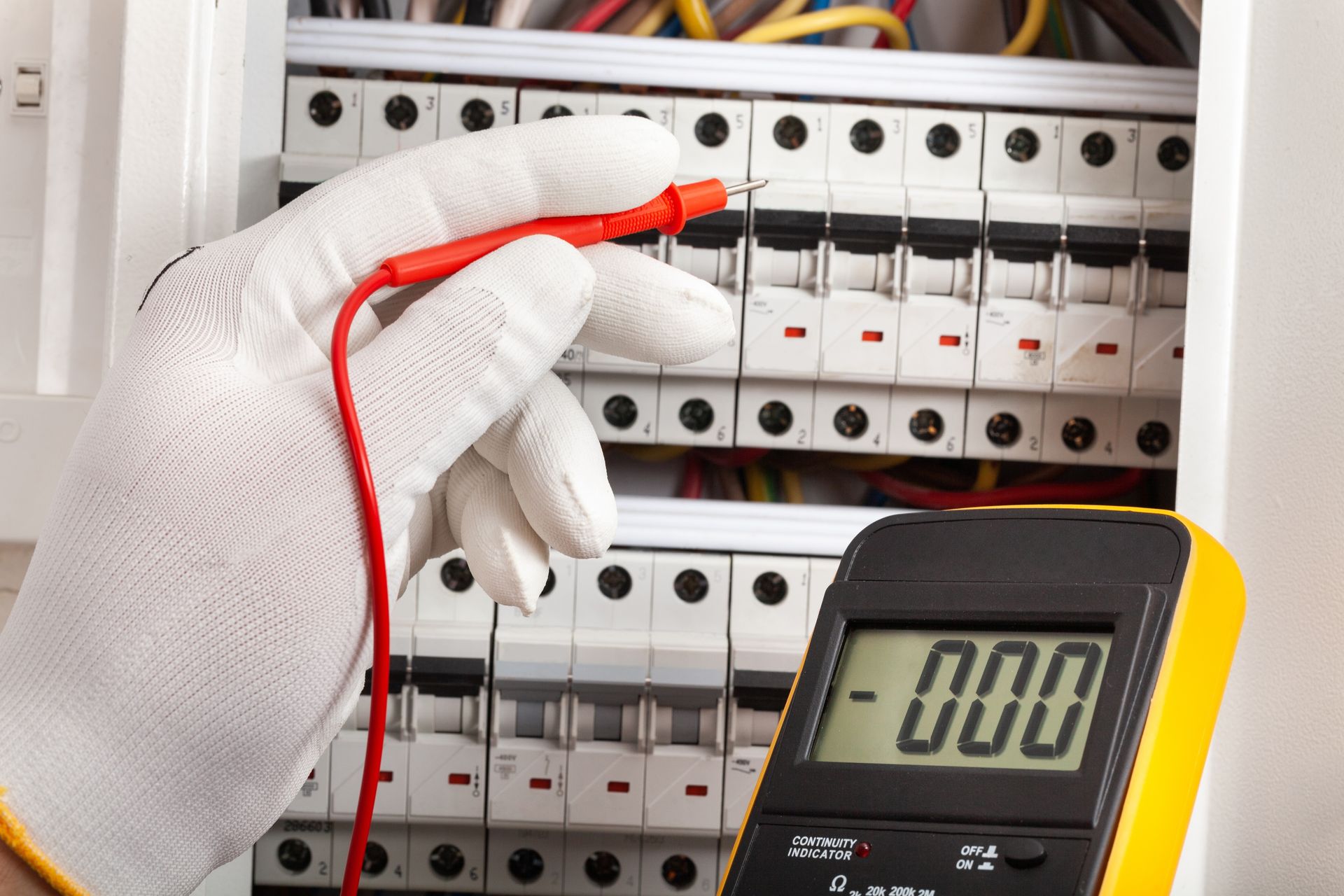 Gloved hand holding a multimeter probe near a circuit breaker panel, indicating a voltage test.