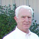 Older man with white hair, smiling, wearing a white shirt, standing outdoors by green bushes.