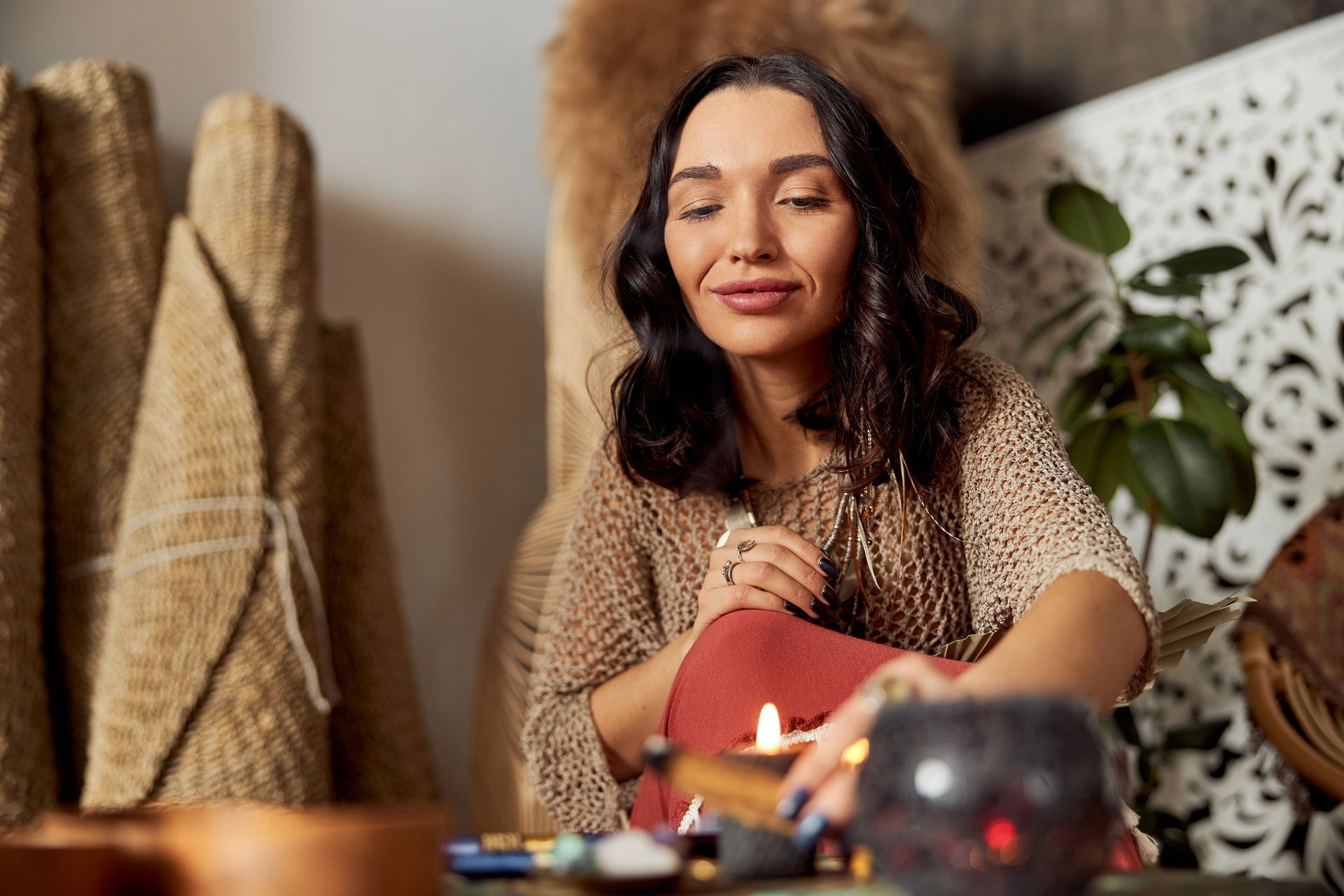 A person wearing a textured beige top sits indoors, thoughtfully holding a burning smudge stick near a small bowl.