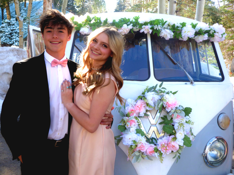 Prom dates are posing in front of the 1967 VW Bus Photo Booth Love Bus, which is grey and white, decorated with pink and white flowers.