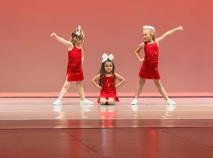 Three little girls in red dresses are dancing on a stage.