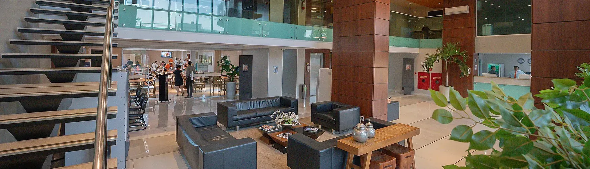 Lobby com escadas, sofás de couro escuro e uma parede de vidro de dois andares. Uma mesa e plantas ocupam o espaço.