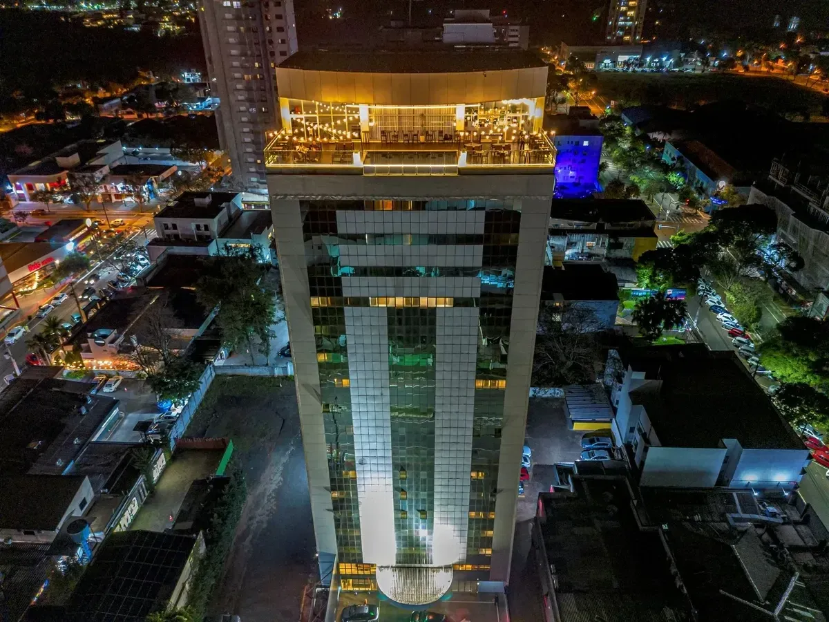 Arranha-céu alto iluminado à noite, com um restaurante no terraço e luzes da cidade ao redor.