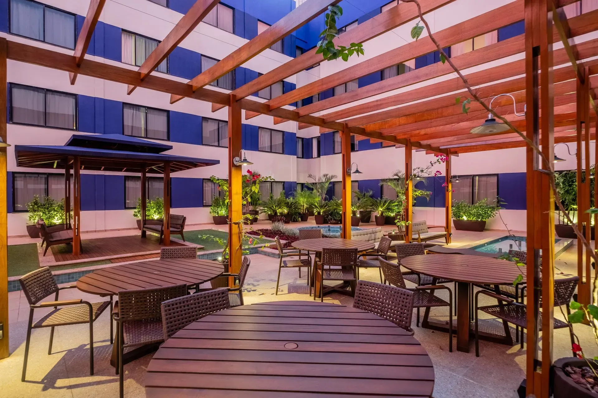 Pátio externo do hotel com mesas, cadeiras e uma pérgola de madeira, com vista para uma pequena piscina e um prédio.