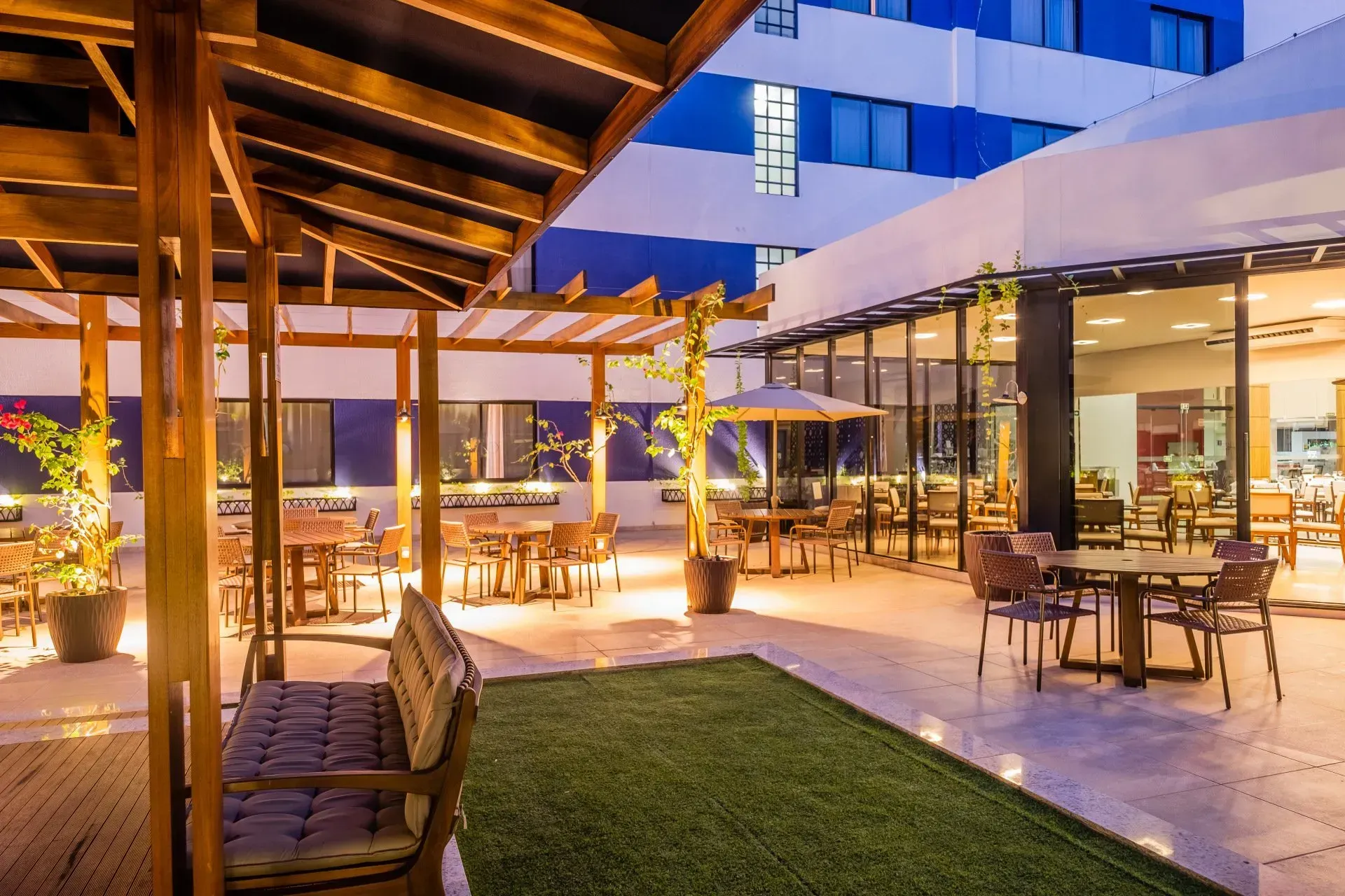 Mesas de restaurante ao ar livre com estruturas de madeira, mesas e um prédio azul/branco ao fundo.