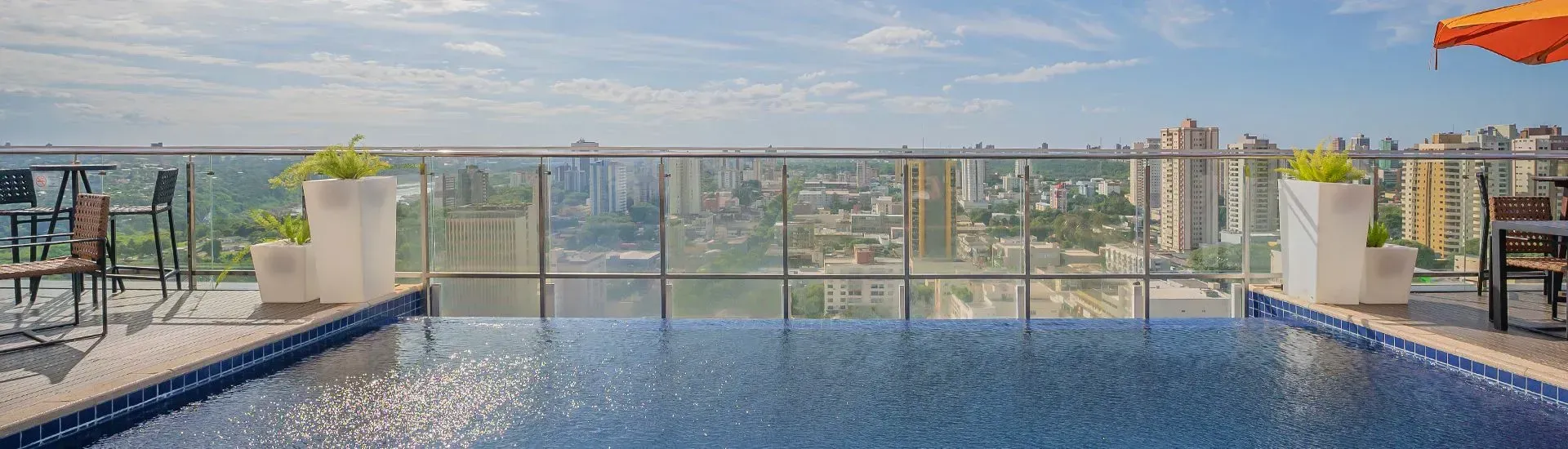 Piscina na cobertura com uma paisagem urbana ao fundo. Vasos brancos decoram as laterais e um guarda-sol laranja é visível.