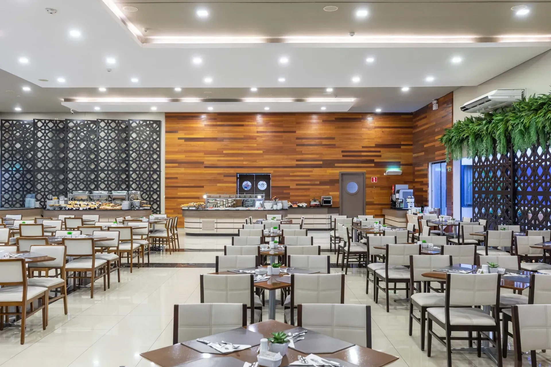 Sala de jantar do hotel com mesas, área de bufê e parede com detalhes em madeira.