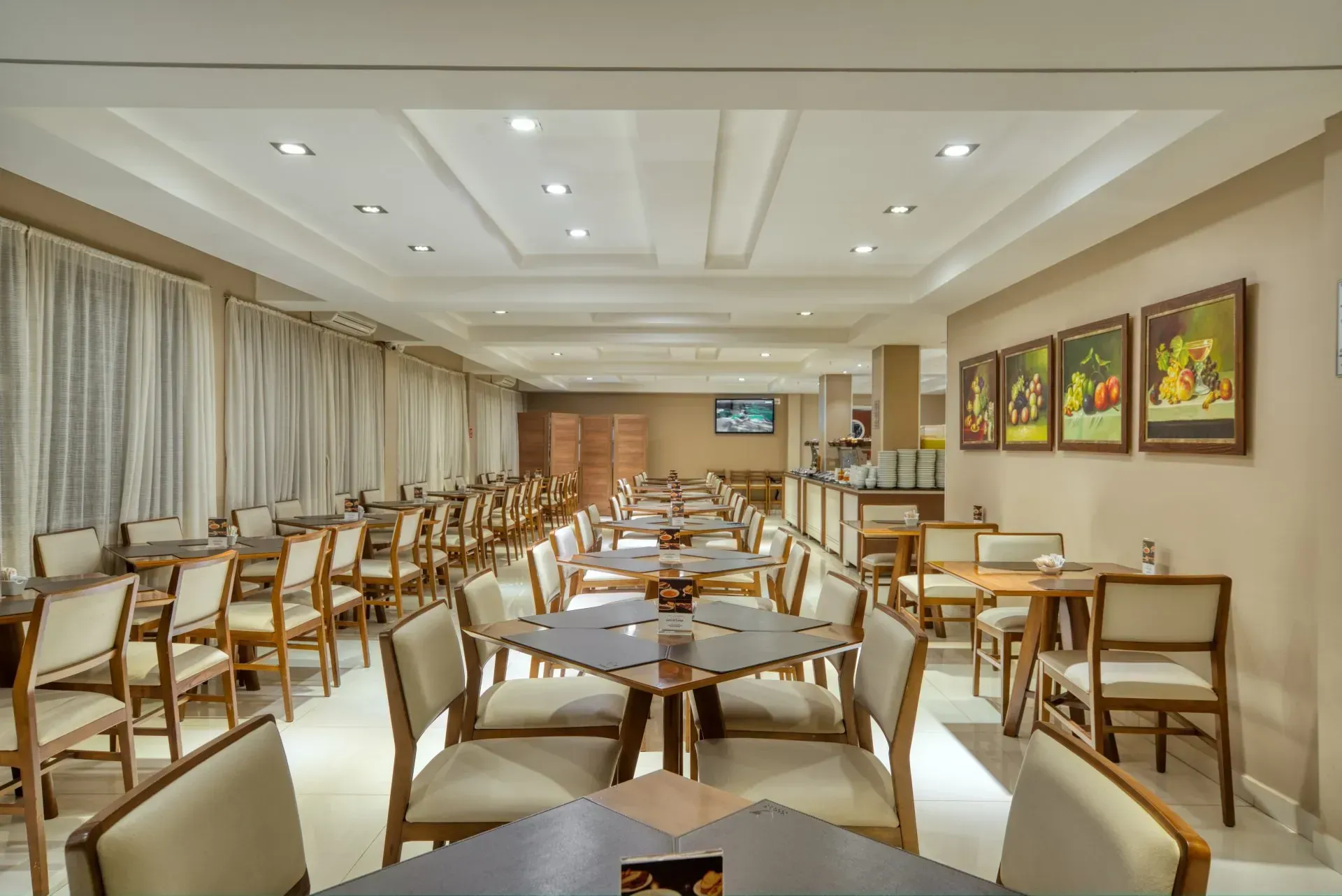 Área de jantar do restaurante com fileiras de mesas e cadeiras, tons neutros e obras de arte na parede.