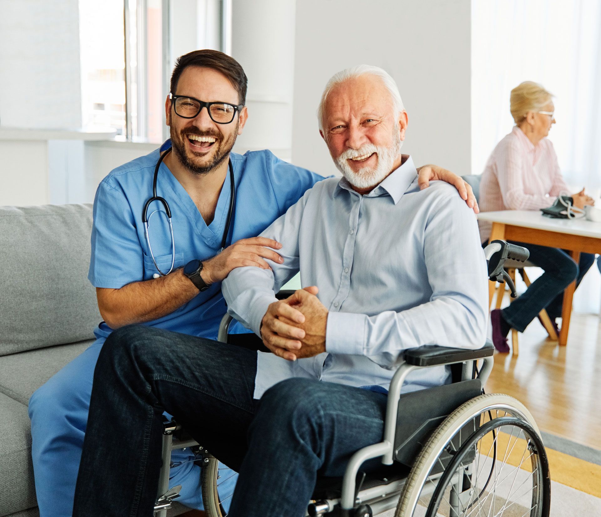 Caregiver in blue scrubs smiles, arm around a smiling senior in a wheelchair. Another person sits at a table.