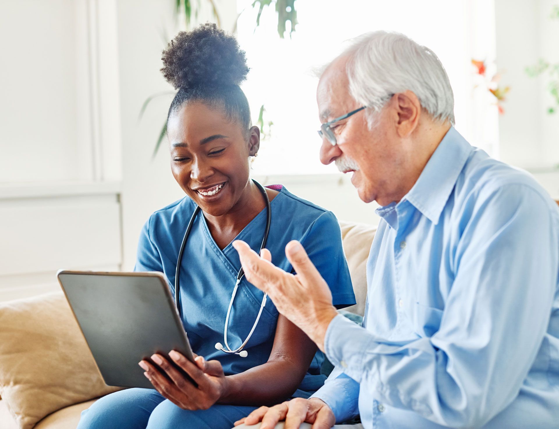 Caregiver shows tablet to an older adult, both smiling; indoors on a couch.