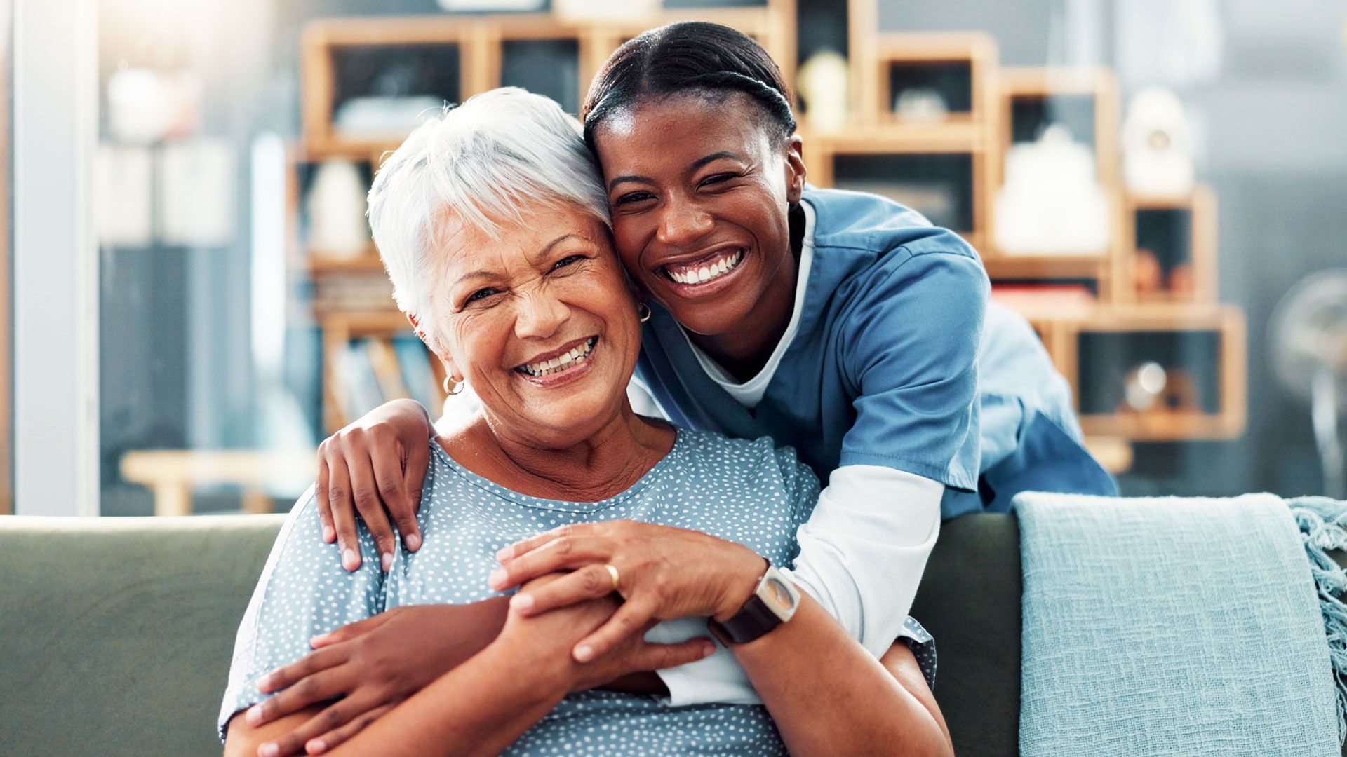 Smiling caregiver hugs senior woman on a sofa.