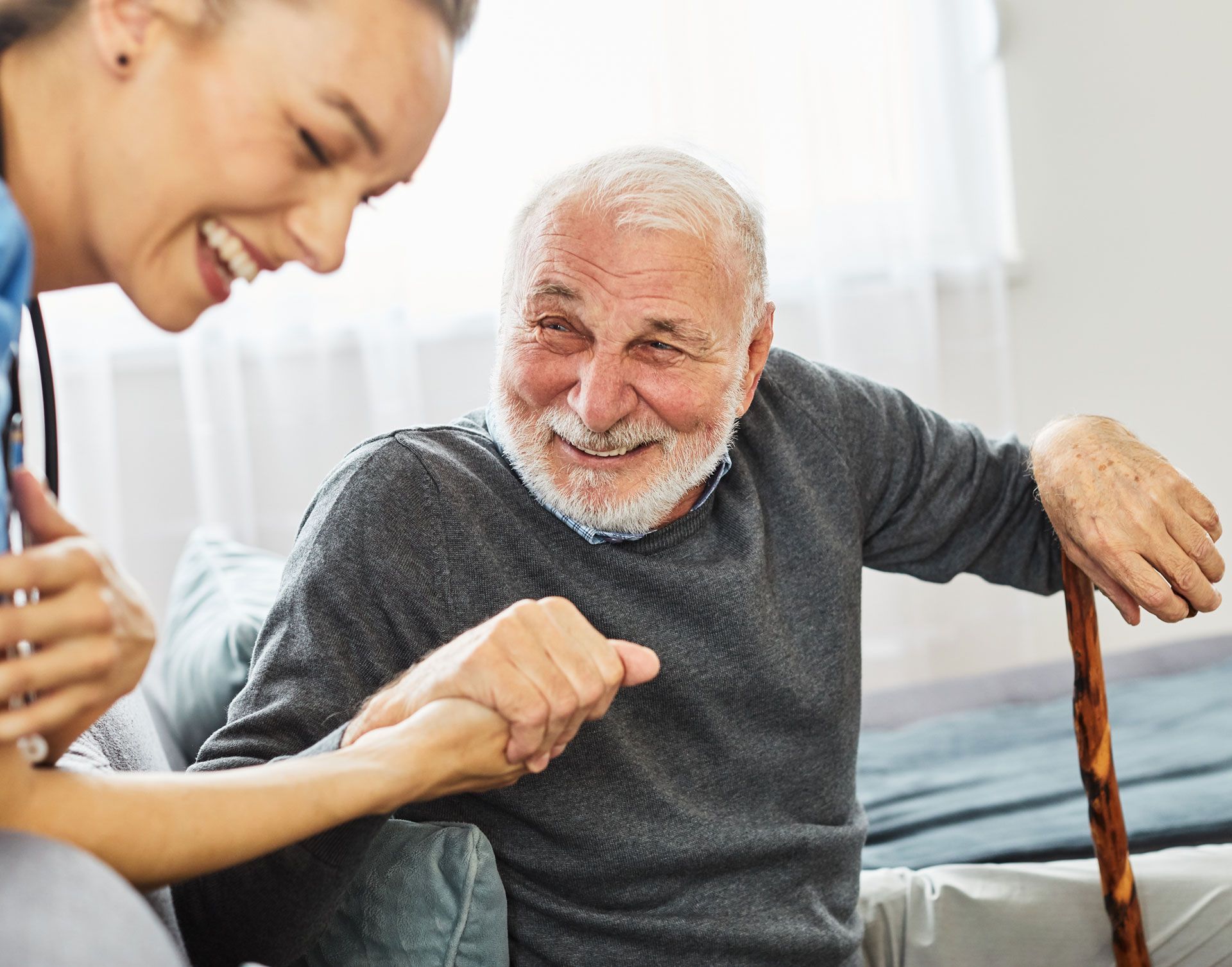 Woman assists a smiling older adult using a cane, indoors. They both appear cheerful.